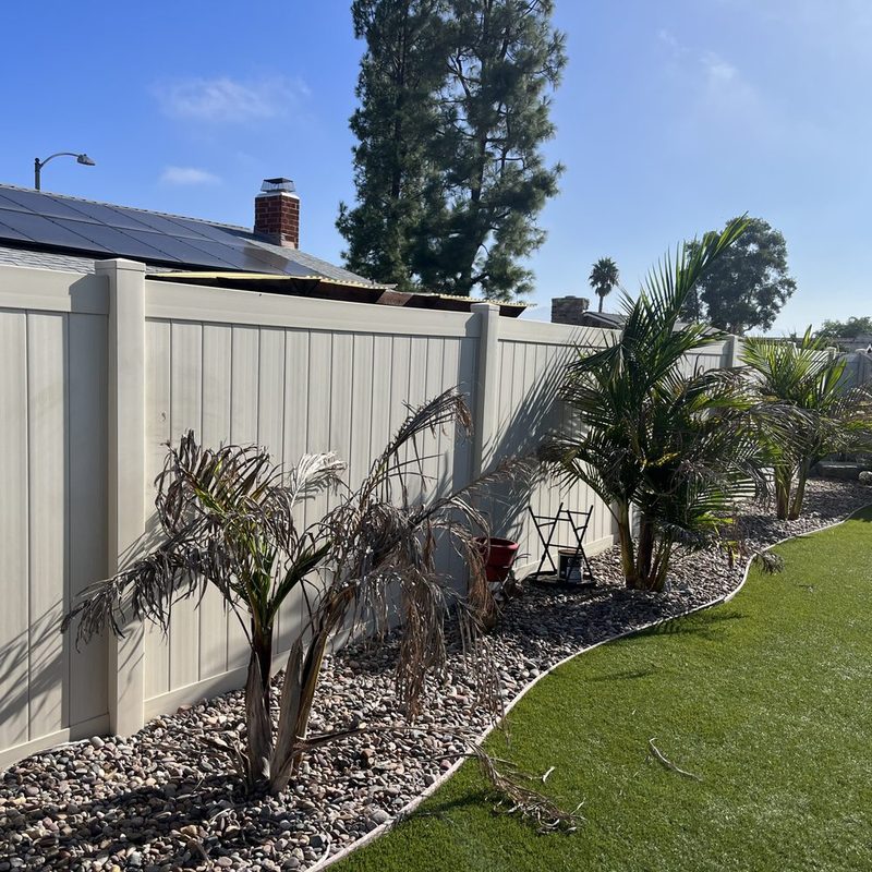 Tan vinyl privacy fence behind a drought-tolerant yard with palms and gravel